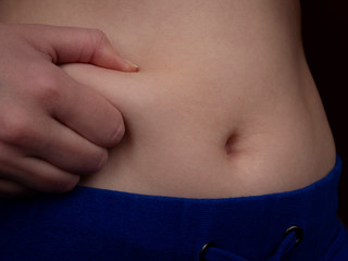 Female belly with excess fat close up. A starting to gain weight young caucasian woman squeezes fat on her waist with her right hand. Unsportsmanlike female body.