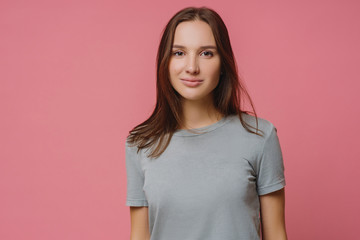 Obraz premium Adorable millennial girl with straight dark hair, wears casual t shirt, looks seriously at camera, has healthy skin, poses over pink background. Pleasant looking woman has talk with someone.