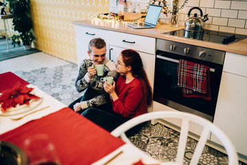 Cheerful young man and woman having hot drinks in kitchen enjoying Christmas holiday at home