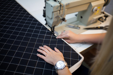 Fototapeta premium Close up view of sewing process. Female hands stitching white fabric on professional manufacturing machine at workplace. Seamstress hands holding textile for dress production. Light blurred background