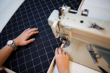 Fototapeta premium Close up view of sewing process. Female hands stitching white fabric on professional manufacturing machine at workplace. Seamstress hands holding textile for dress production. Light blurred background