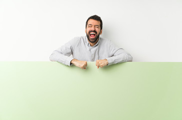 Young handsome man with beard holding a big green empty placard shouting to the front with mouth wide open