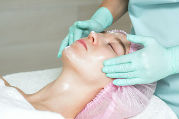 Cosmetologist's hands in gloves applying a white cream to the woman face in the spa beauty salon...