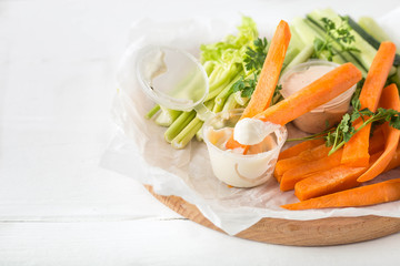 Diet raw sticks of cucumber, celery, carrots with yogurt white sauce on a board with parchment on a white background. Image with horizontal orientation and copy space