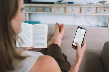 Female on couch surfing smartphone while reading book at home