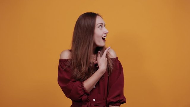 Surprised caucasian woman in maroon dress isolated on orange background in studio pointing finger aside, looking at camera. People sincere emotions, lifestyle concept.