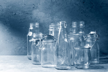 Variety of different shape empty opened glass bottles with and without lids standing on grey table with wall as background. Color of the year 2020 classic blue toned