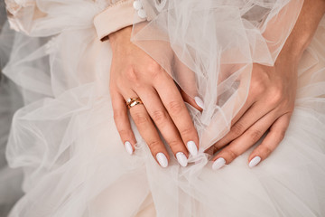 close-up of the hand of a bride who sits clasped hands in a delicately pink dress with a ring on...