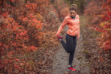 Young attractive sport woman in sportswear doing stretching leg in autumn forest with reddened leaves of trees. Warm up before running. Healthy lifestyle concept