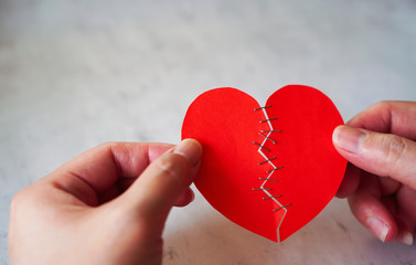 Fototapeta premium Hands of woman hold and heal broken heart made from red paper by stapler ,on white concrete background, represent Love , Care , healing heart and recover from pain of heartbreak between couple