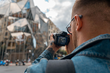 Attractive Man Focusing Photo Camera On Modern Building. Cityscape Background Is Blurred. Blogger and Traveler. European holidays