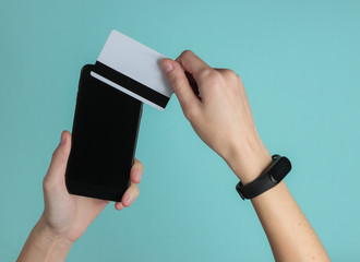 Hand with smart bracelet holding bank card and smartphone on blue background. Cashless payment, online shopping. Top view.