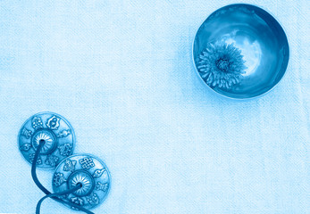 A small singing bowl stands in the upper right corner filled with water with floating chrysanthemum inside. A small singing bowl and tingsha stand on background of organic cotton. Top view.