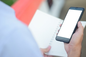 close up hand holding white screen smart phone and book for plan work at outdoor over blured background.