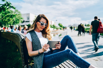 Cute stylish hipster girl in cool sunglasses spending free time at urban setting sitting on wooden bench and reading news in webpage.Good looking female blogger making publication in personal website