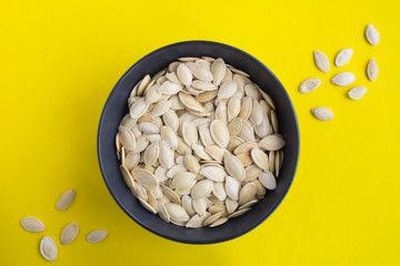 Pumpkin seeds in the black  bowl in the center of the yellow  background. Top view. Copy space.
