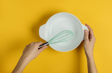 Female hands holding empty ceramic bowl and whisk on a yellow background. Top view