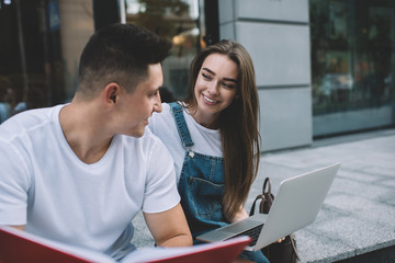 Positive male and female students in casual wear sitting at urban setting and discussing details of university course work using modern laptop computer for research information, concept of education