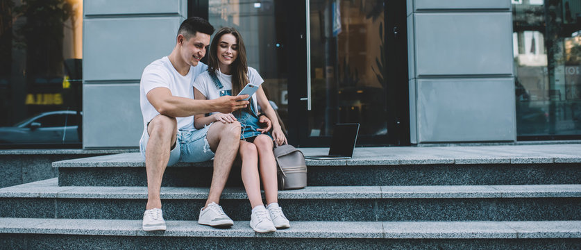 Cheerful hipster guys sitting at city stairs connecting to public internet for watching online video vlog in social networks, positive couple in love enjoying leisure time togetherness for chatting