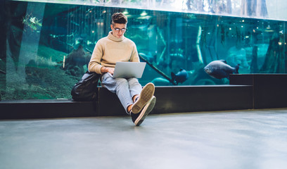 Young man using laptop and sitting with legs crossed near aquarium