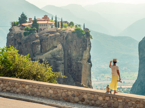 Young woman with white dress and large hat standing on wall in front of greece meteor mountains, monastery and village in the background