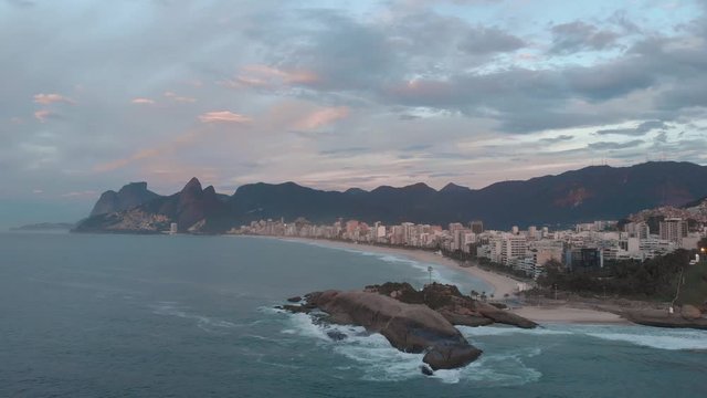 Aerial tilt down cityscape of Rio de Janeiro, Brazil, of Ipanema beach with the Arpoador rock in the foreground and Two Brothers mountain and Gavea in the background at sunrise with orange clouds 