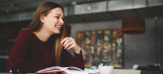 Attractive skilled happy female student sitting at cozy cafe smiling carefree at break, cheerful hipster girl laughing at funny stories from textbook enjoying doing homework at college library