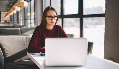 Serious hipster girl 20 years old in spectacles looking forward while sitting indoors in cozy...