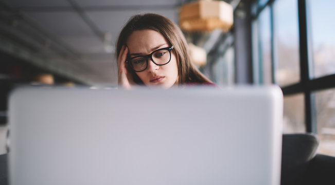 Bored pensive hipster girl in eyeglasses watching webinar online concentrated while sitting at university campus, tired female student studying online indoors using netbook for exam preparation