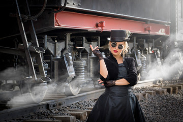Steampunk girl in a black dress and hat near an old steam locomotive and large iron wheels. Blond beauty. Vintage portrait of the last century, retro journey.