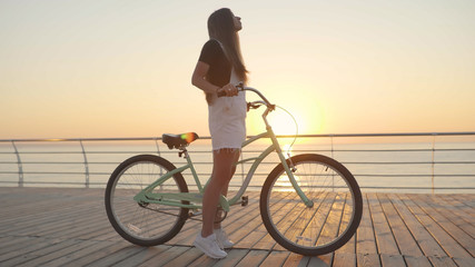 Obraz premium Young beautiful woman with a bicycle is having a good time at the sea at sunset or sunrise 