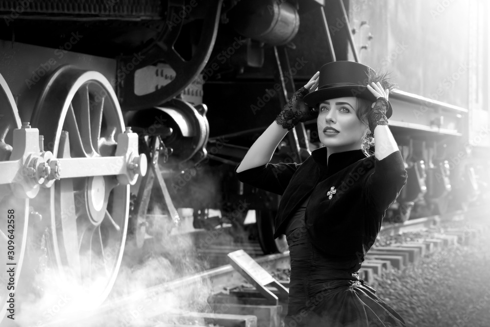 Wall mural Beautiful girl in a black dress and hat near an old steam locomotive and big iron wheels. Blond beauty. Vintage portrait of the last century, retro journey.