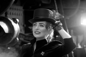 Beautiful girl in a black dress and hat near an old steam locomotive and big iron wheels. Blond beauty. Vintage portrait of the last century, retro journey.
