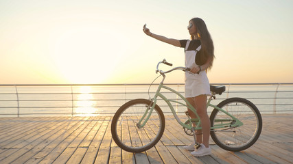 Fototapeta premium Young attractive woman uses a smartphone and riding vintage bike near the sea during sunrise or sunset