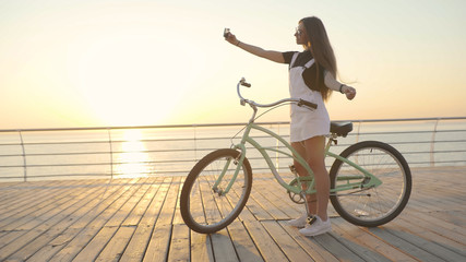 Young attractive woman uses a smartphone and riding vintage bike near the sea during sunrise or sunset