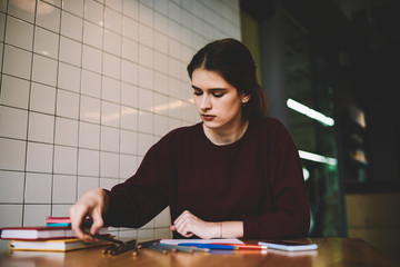 Attractive skilled hipster girl choosing pencil for drawing sketches while sitting at academy library,  thoughtful woman learning and preparing for exam at university campus while waiting friend