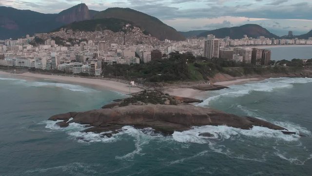Slow aerial tilt down showing the Arpoador rock in Ipanema, Rio de Janeiro, with waves crashing into it at sunrise with the cityscape including shantytown on a hill in front of the Corcovado mountain