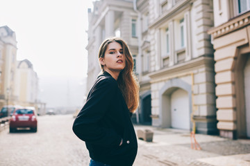 Fototapeta premium Portrait of beautiful female student in black jacket enjoying free time while walking across city and looking at camera, trendy dressed street style and fashionable hipster girl strolling outdoors