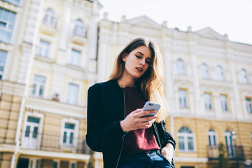 Hipster girl sending text messages and searching information about touristic route via cellphone outdoors, beautiful trendy woman chatting with friends online in social networks using modern telephone