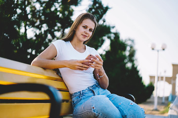 Portrait with positive hipster girl holding mobile phone in hand looking at camera resting leisure in park, carefree female blogger using cellular for chatting while browsing 4g internet outdoors