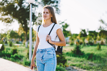 Smiling teenage caucasian woman resting on summer leisure and strolling across street on urban setting, young positive hipster girl 20s in jeans walking outdoors during free time on weekend