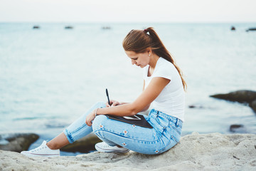 Charming teenage hipster girl in denim jeans writing interesting review about travel in notebook outdoors, young fashionable beautiful woman enjoying drawing sketch nature near calm lake surface