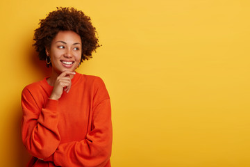 Happy moment. Positive curly girl holds chin, looks aside, sees something pleasant, has broad smile, dressed in casual jumper, stands against yellow background, blank space aside. Lucky black female