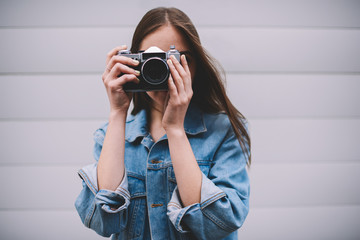 Portrait of talented hipster girl posing near wall while making pictures using obsolete vintage camera, young professional female photographer in leans jacket standing outdoor photographing