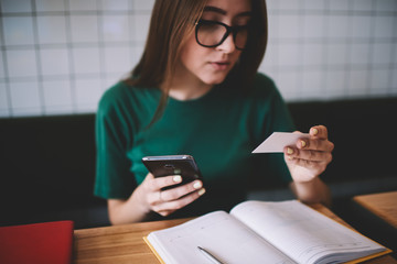 Serious hipster girl looking at paper reading mobile number and dialing friend using modern smartphone, young female in spectacles  student holding telephone in hand while contact service operator