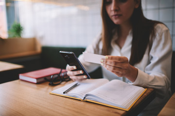 Young hipster girl 20s holding smartphone in hand and looking at business card for dialing colleague for discussing meeting, female student writing email via modern mobile phone while sitting indoors