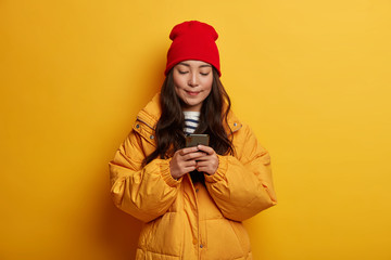 Fashionable Asian woman with dark long hair, wears winter outfit, texts on mobile phone, browses website online, being obsessed with modern technologies, isolated on yellow wall. Communication.