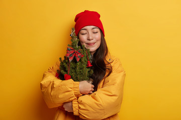 Portrait of attractive woman keeps eyes closed, embraces decorated fir tree, dressed in winter jacket, imagines nice celebration of holiday, isolated over yellow wear. People, winter concept