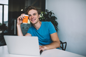 Portrait of cheerful hipster guy with coffee to go sitting at desktop with modern netbook for...