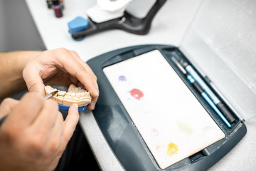 Dental technician coloring dental prosthesis with a paint brush at the laboratory, close-up view. Concept of implantats producing